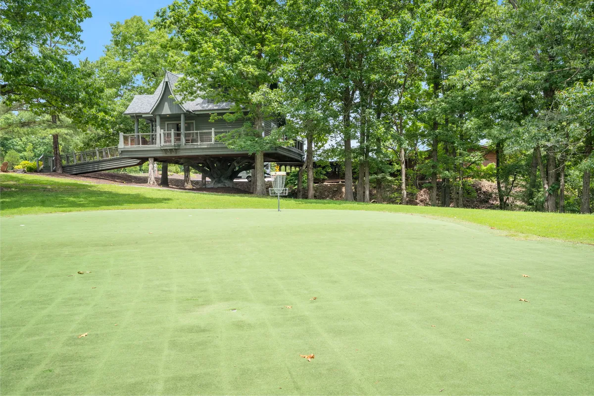 This image features a house on stilts overlooking a green lawn, with a golf basket in the foreground and lush trees surrounding the scene. The setting evokes outdoor activity and relaxation, capturing the essence of disc golf and nature-based recreation.