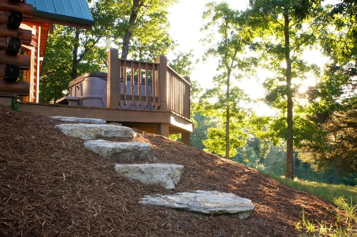 A private hot tub sits on a wooden deck, surrounded by trees and accessed by stone steps. The setting suggests peaceful relaxation after a day filled with outdoor activities.
