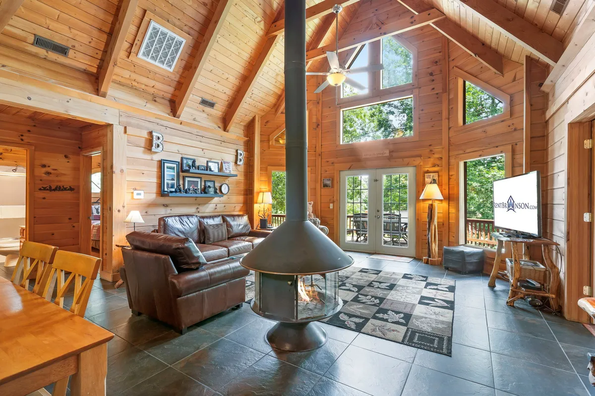 A soaring living room with vaulted wooden ceilings, a central fireplace, comfortable leather couches, a large dining table, and expansive windows offering beautiful views. The layout encourages relaxation, conversation, and shared moments.