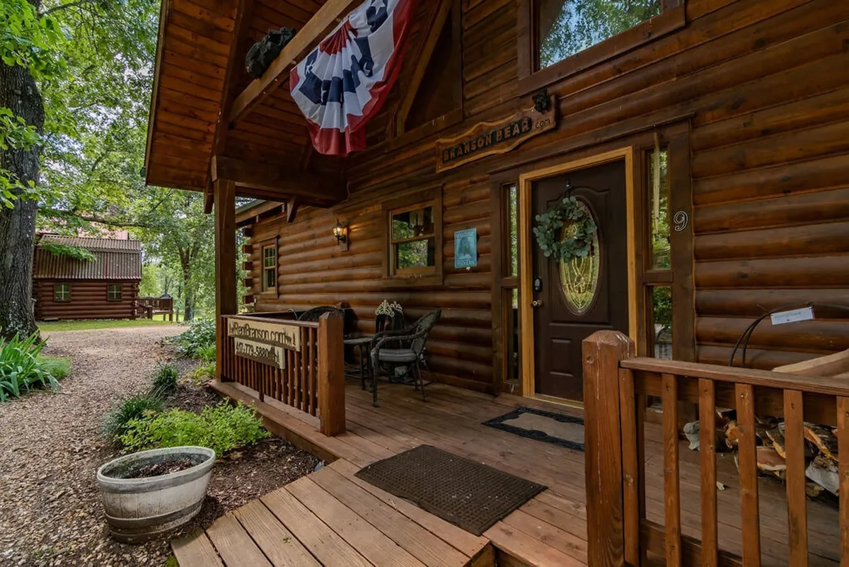 A cozy exterior shot of a log cabin featuring a front porch, decorative wreath, and festive flag bunting. The porch overlooks inviting natural surroundings and suggests a welcoming spot to relax after adventures.
