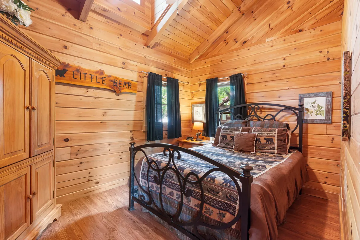 A queen bedroom with rustic wooden walls, a comfortable bed covered in patterned bedding, a 'Little Bear' decorative sign above the headboard, and soft natural light streaming through the window. The setting feels cozy and peaceful.