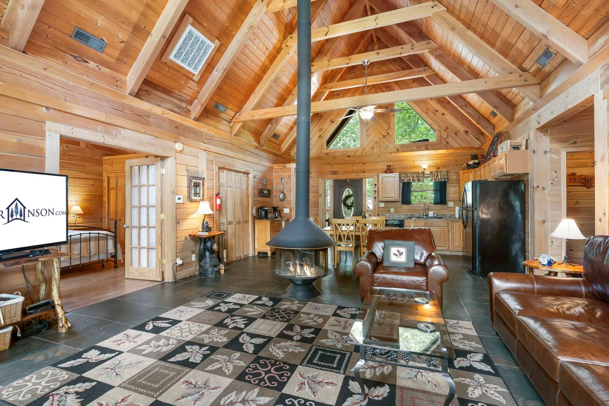 A warmly lit open-concept living space featuring a central circular fireplace, plush leather sofa, spacious dining area, and large windows that flood the room with natural light. The kitchen is visible in the background, seamlessly connected to the rest of the gathering space.
