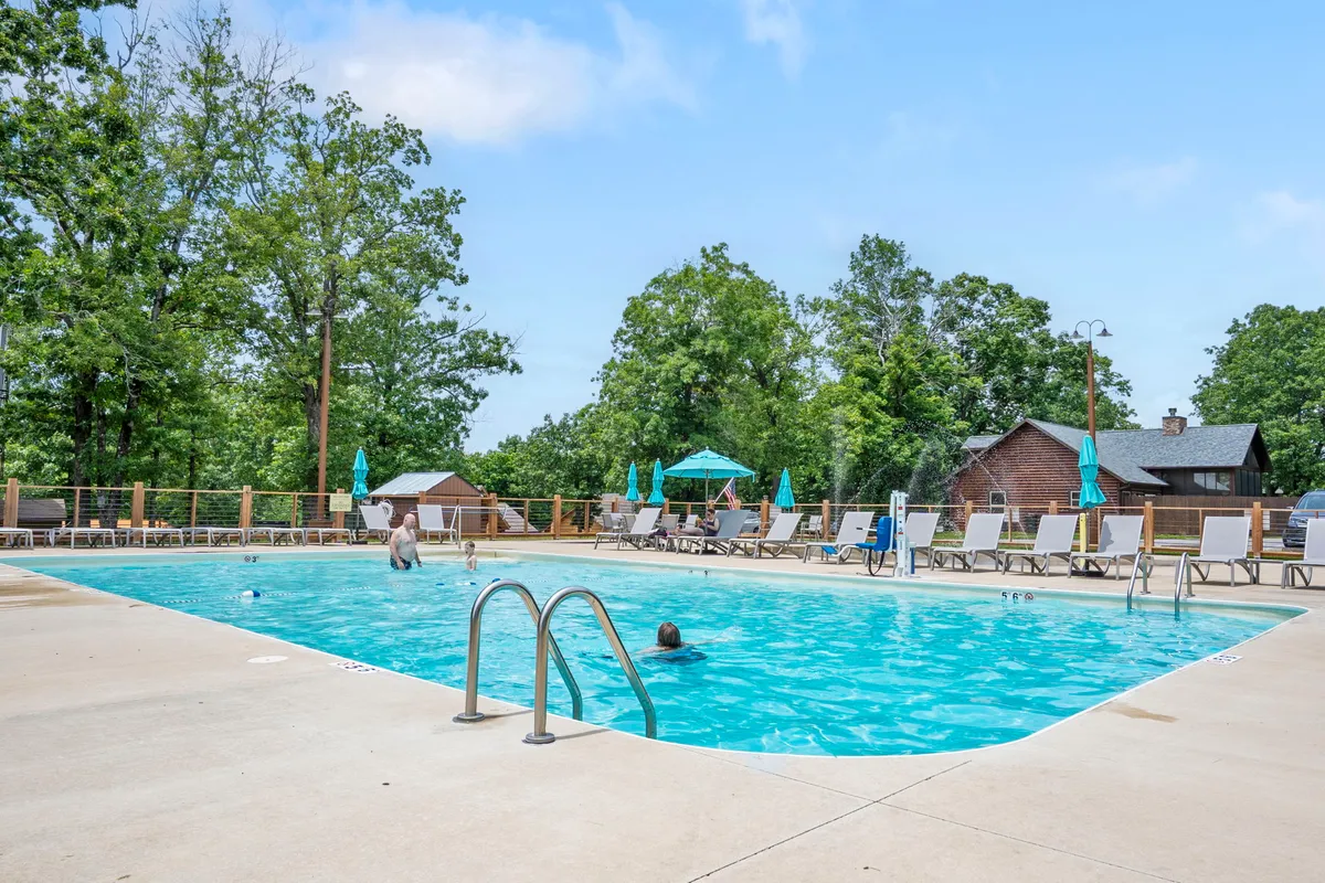 A sun-drenched swimming pool with people swimming, surrounded by lounge chairs, umbrellas, and trees, creates a lively, fun-filled resort atmosphere ideal for family enjoyment and relaxation.