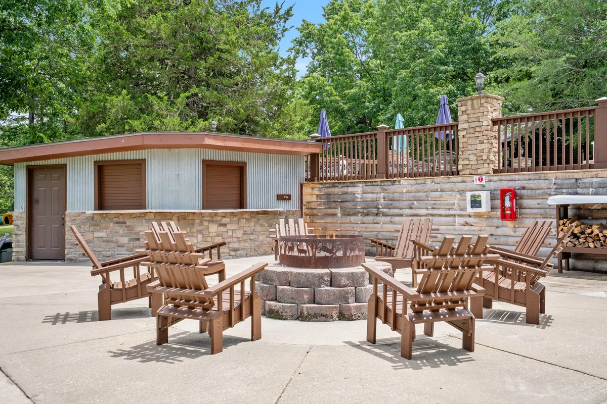 A cozy communal fire pit encircled by Adirondack chairs sits near a wooden deck, set against a backdrop of lush green trees. The scene evokes a sense of togetherness, perfect for stargazing, storytelling, or enjoying s'mores by the fire.