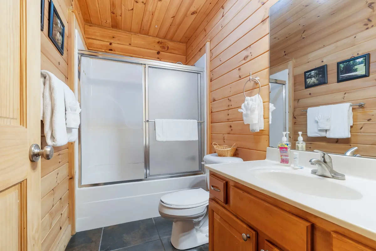 This shot captures a well-appointed hallway bath with wooden paneling, a spacious shower area, sink with mirror, toilet, and neatly arranged towels. The space feels clean, functional, and inviting.