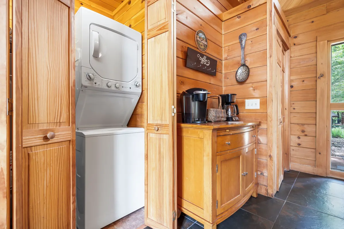 A bright, inviting laundry area with a modern washer and dryer set, wooden cabinetry, a coffee station, and natural light streaming in from a window. The space feels organized and practical, with essentials for daily routines easily accessible.