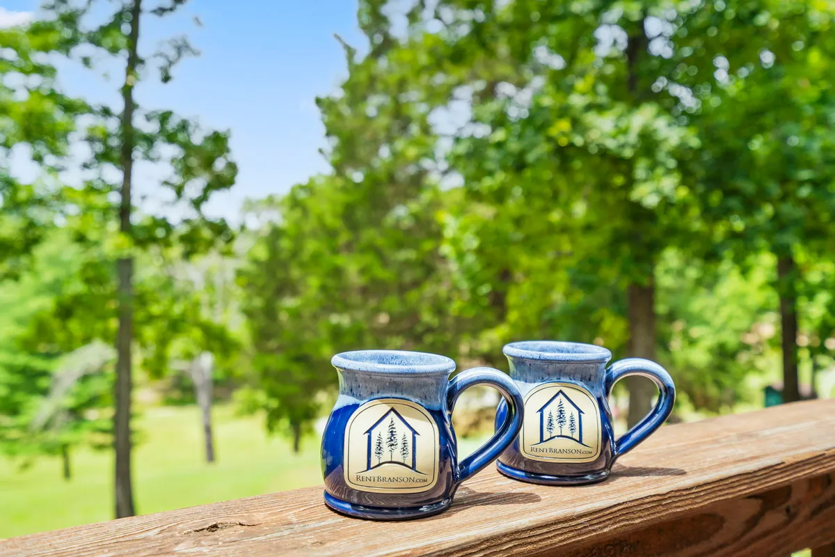 This image captures two blue mugs set on a wooden deck rail, with a backdrop of lush green trees. The scene evokes the tranquility of enjoying a quiet morning coffee outdoors, immersed in nature.