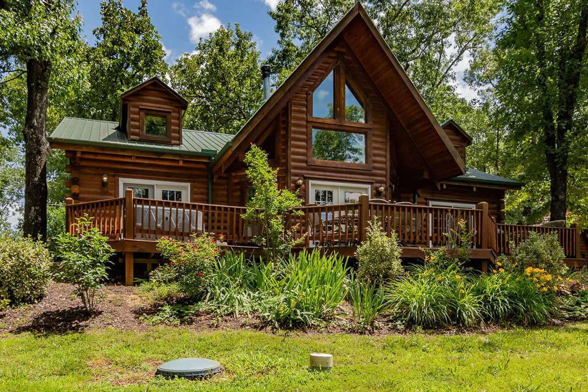 The exterior of a log cabin with a spacious deck, large windows, and a lush garden setting. The scene is inviting, with plenty of space around the cabin for parking vehicles or small boats, as mentioned in the features list.