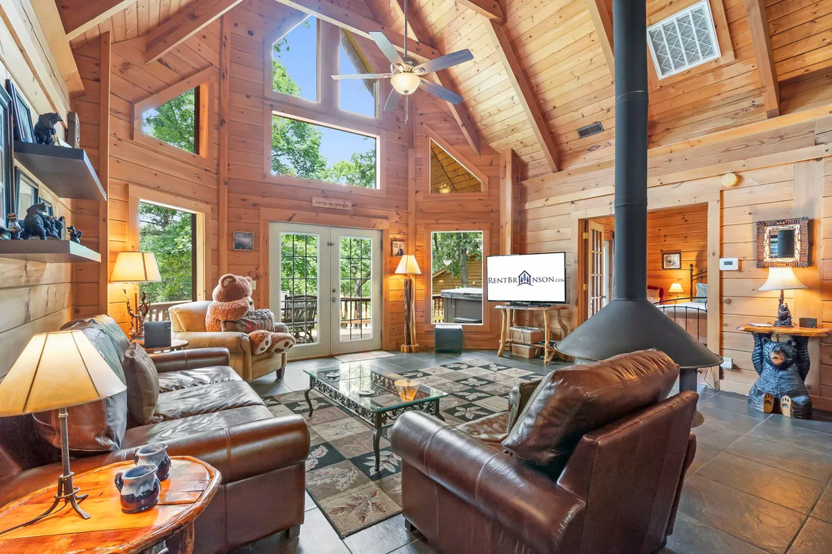 The hero image showcases a spacious cabin living room with soaring vaulted ceilings, exposed wooden beams, a stunning stone fireplace, floor-to-ceiling windows offering views of the surrounding woods, and plush, inviting seating arranged for relaxation and socializing. Sunlight streams through the windows, highlighting the natural wood textures and creating a cozy, welcoming atmosphere.
