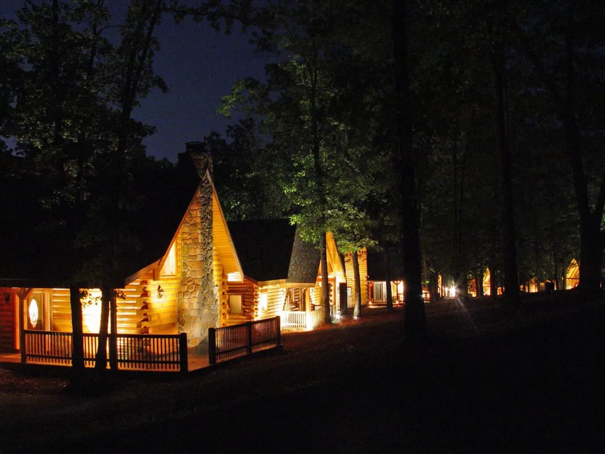 A tranquil night scene showing warmly lit log cabins, silhouetted trees, and an inviting atmosphere perfect for stargazing or evening strolls. The overall mood is peaceful, cozy, and romantic.