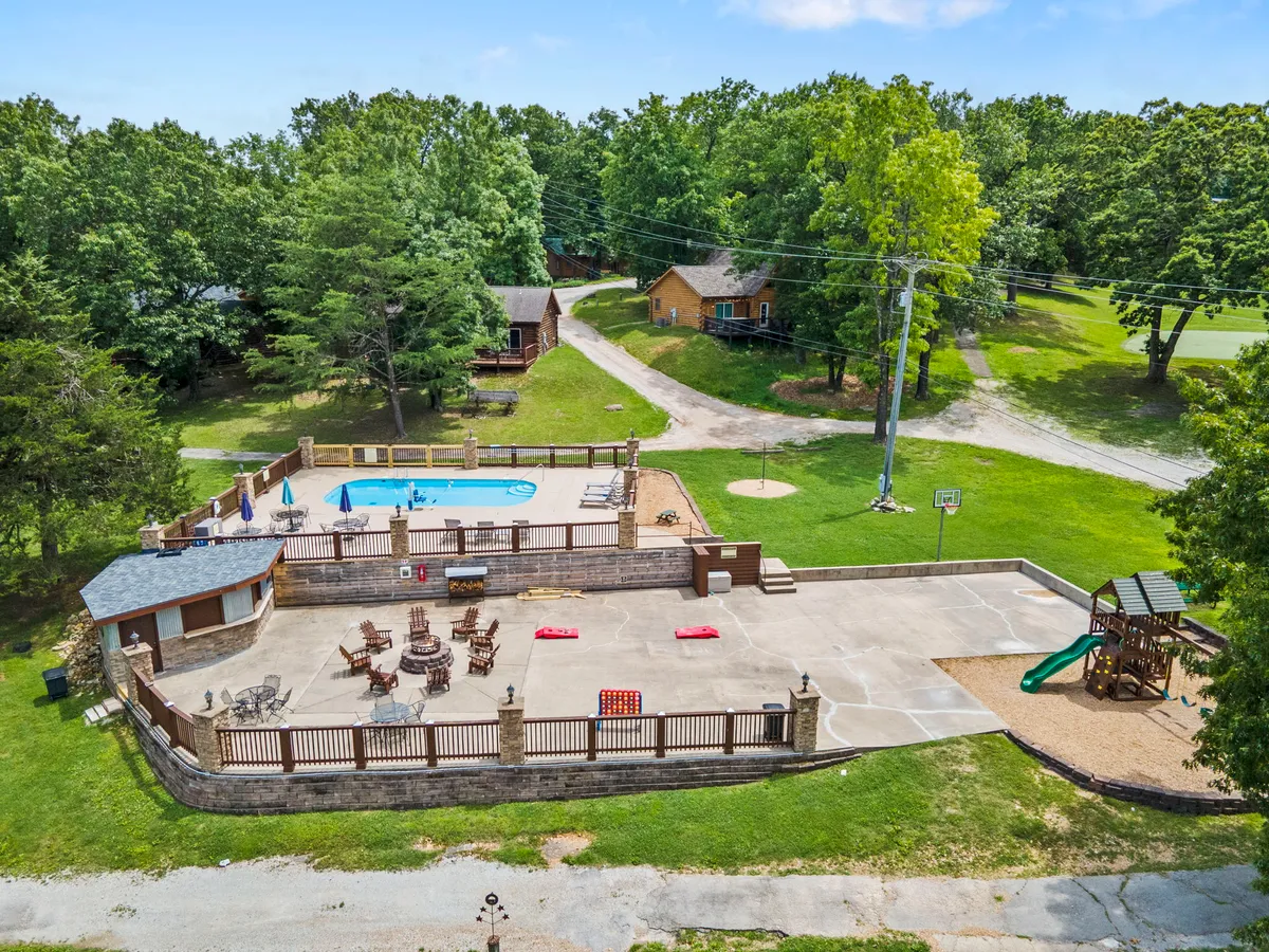 A lively exterior scene showcasing a swimming pool surrounded by lounge chairs and a playground area, with a basketball hoop and cabins visible in the background. Children and families can be imagined basking in the sun, splashing in the pool, or playing on the playground, all set within a resort environment.