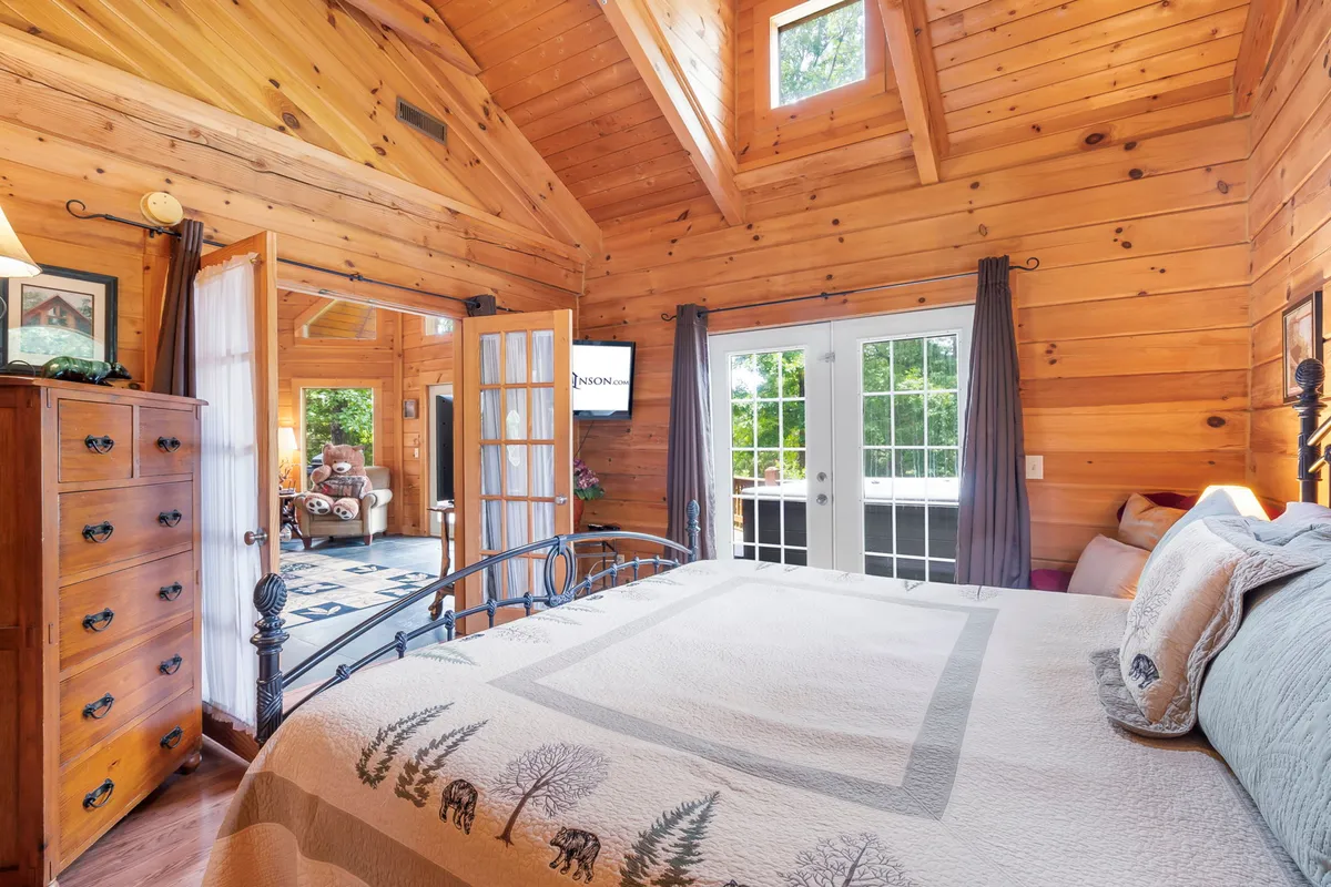 This image showcases a cozy bedroom with wooden walls, a quilted bedspread, and French doors that open to the outside, letting in plenty of natural light. A decorative lamp sits on a bedside table, and the space is inviting and tranquil.