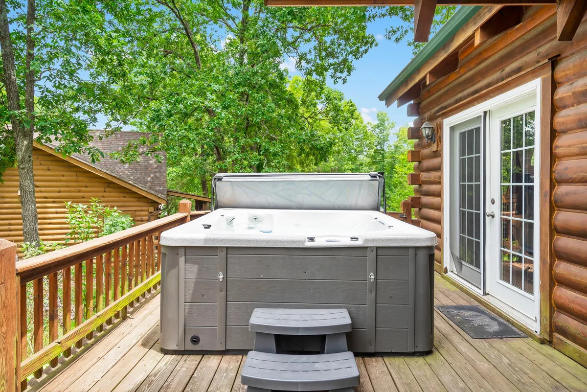 A private wooden deck featuring a bubbling hot tub, with trees and greenery surrounding the area. Sliding glass doors open from the cabin, creating a seamless indoor-outdoor connection, and the setting is perfect for relaxation under the stars.